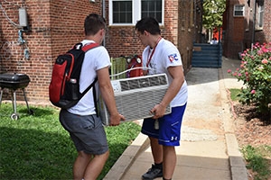 Two students carry an air conditioner unit into a building.