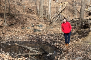 Person in a jacket stands in the woods next to a stream