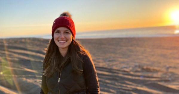 Alumna Sarah Diamond stands outside at the beach at sunset while wearing a red hat and black zip up jacket.