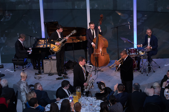 Well-dressed musicians play the cello, guitar, saxophone, piano, and trumpet onstage.