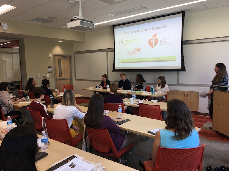 People sit in a class room listening to a panel of four professionals. A presentation slide reads 