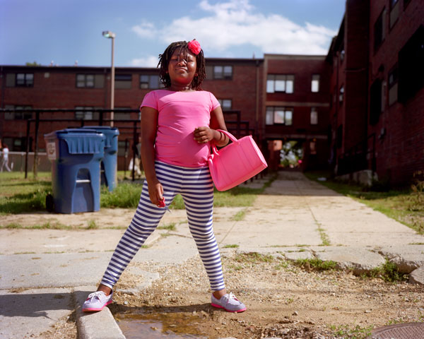 Photo of a young girl in a bright pink shirt and striped leggings standing in the sunshine astride a curb in front of some recycling bins and a brick building. She holds a pink purse and a pair of red scissors.