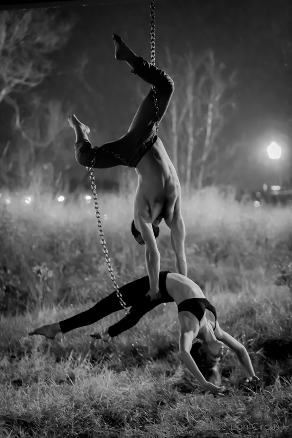 Black and white photo of a man suspended upside-down by a chain on his waist using only his hands to hold a woman who leans back parallel to the ground. They are in a grassy field. A streetlight glows in the right upper corner.