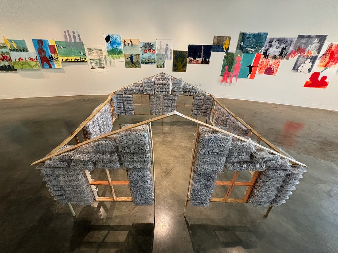 A model of a house framed with wood and insulated with recycled paper and plastic. In the background, assorted prints from The Human Flood.