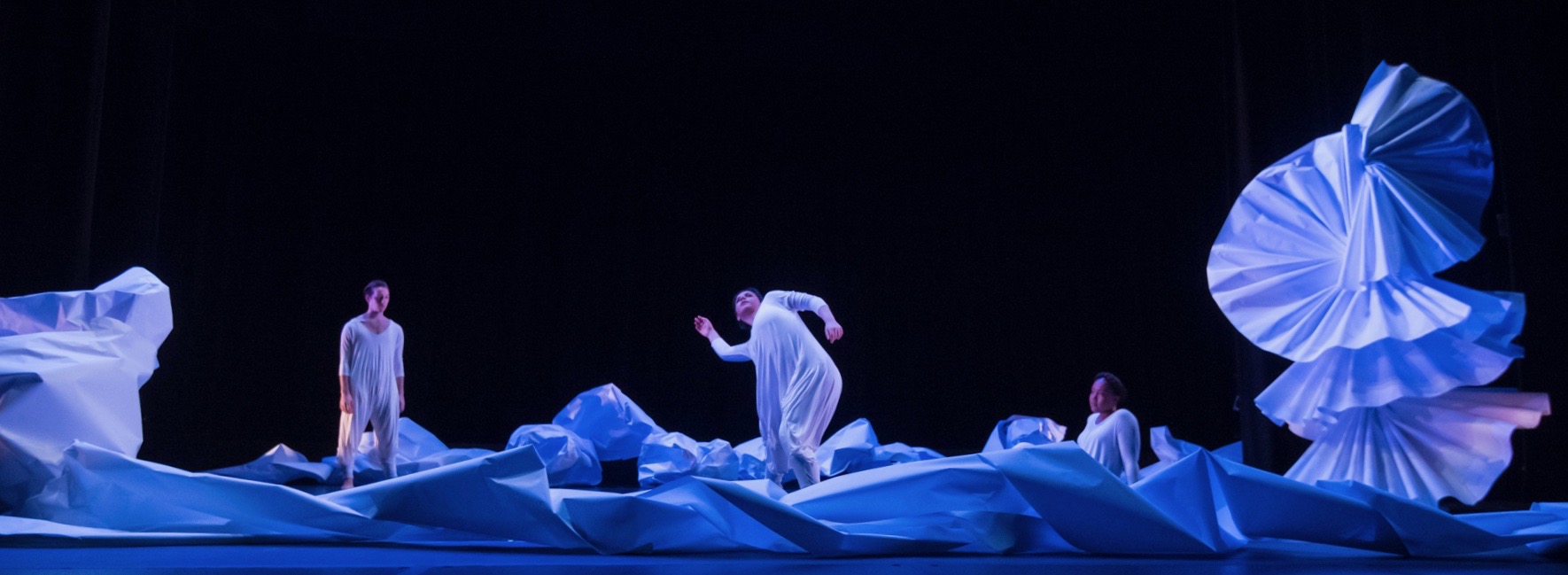 Dancers watch another dancer reach toward the sky, while sitting among a sea of paper and a large paper sculpture