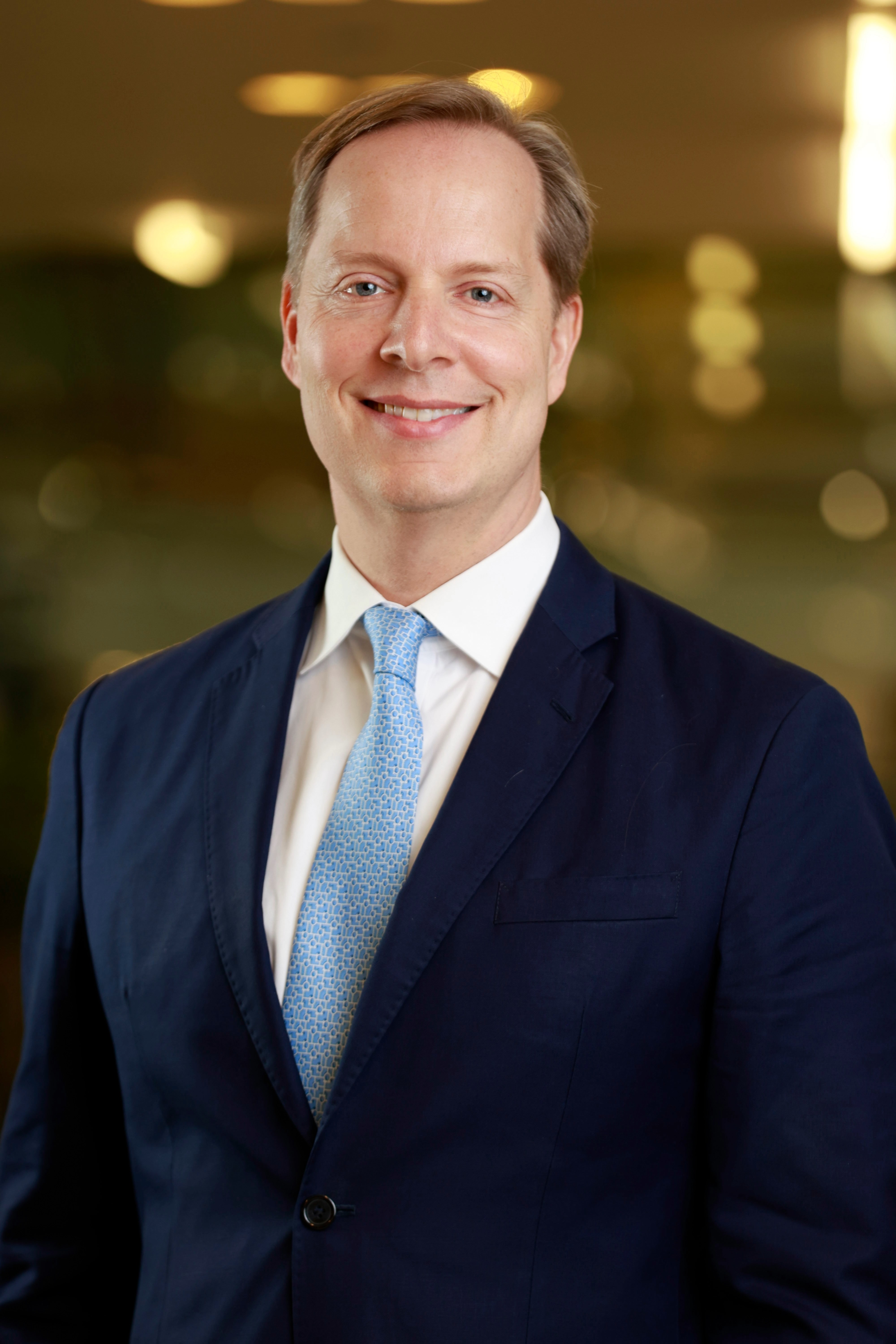a white man with blond hair wearing a blue suit and tie smiles at the camera