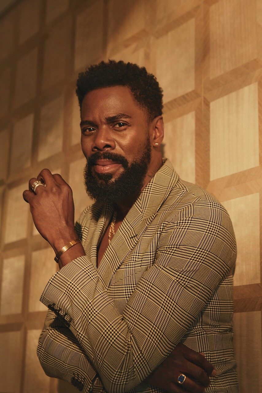 a man in a blazer leans up against a wall and looks at the camera