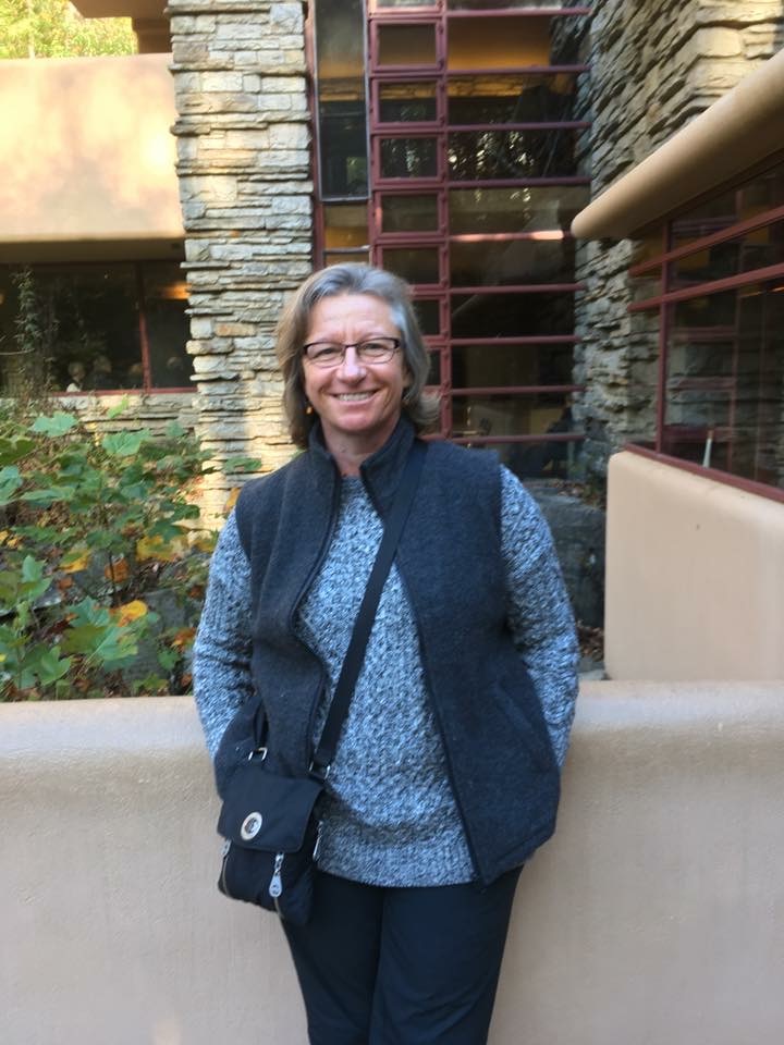 A photo of Prof Schaeff standing outside of a building. She has shoulder length hair and is wearing a blue sweater with a dark blue vest