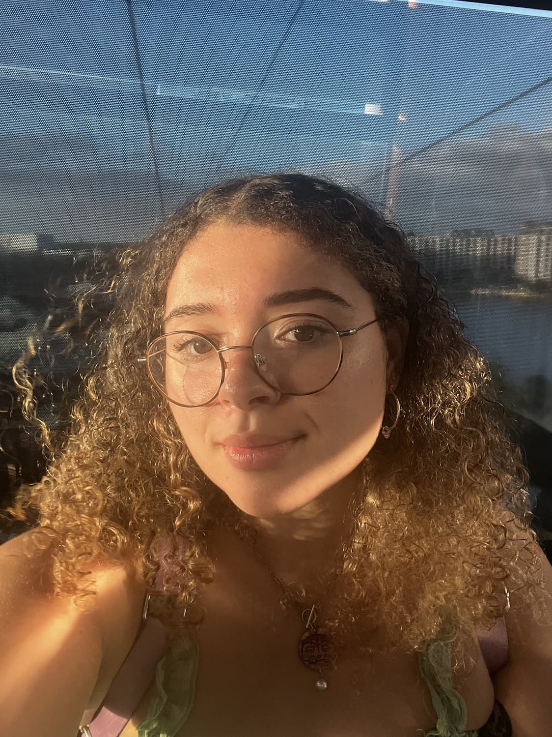 Cheyenne, a young person with medium-dark skin, glasses and curly brown hair in front of a city and a body of water.