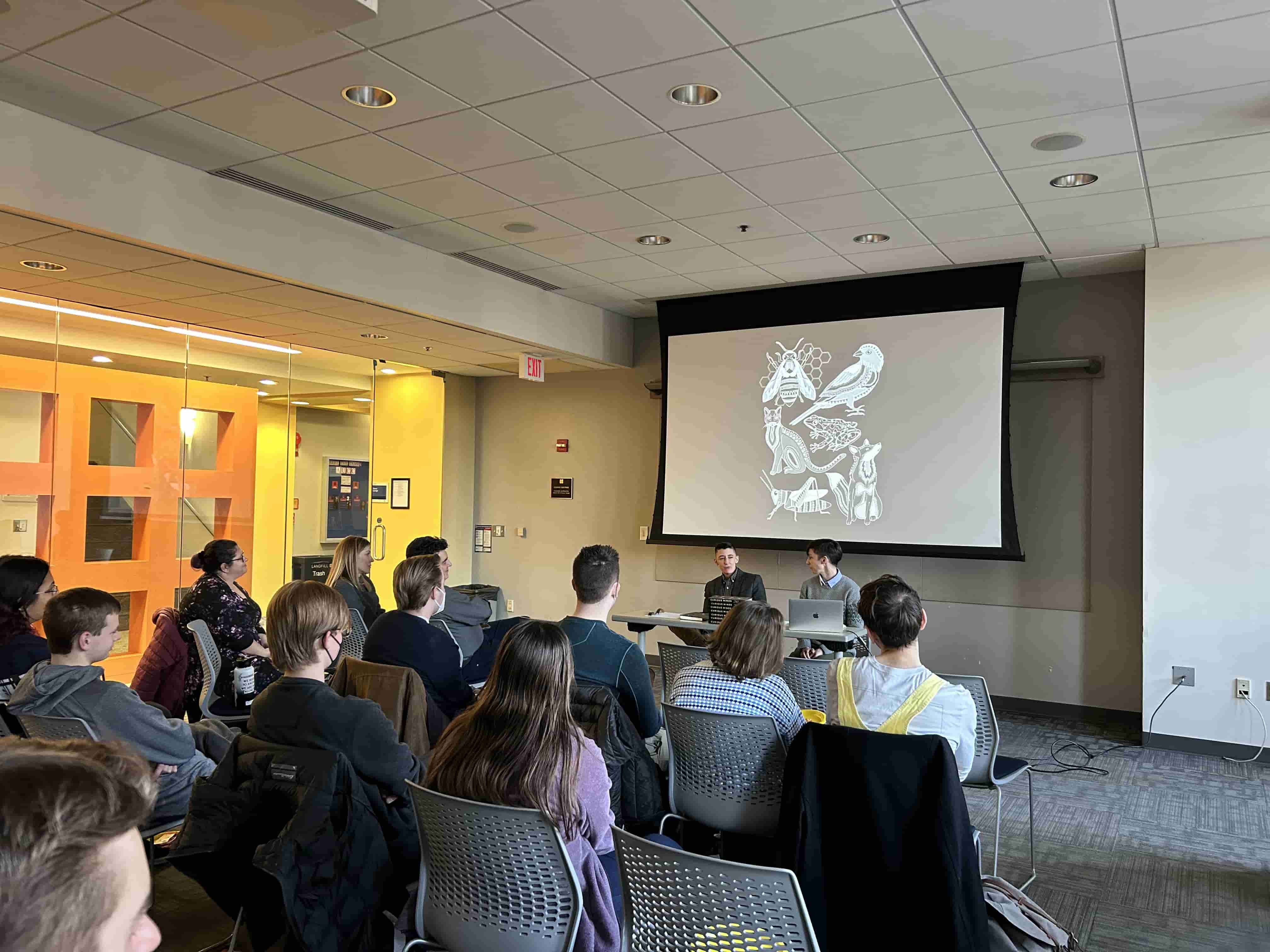Group of students sitting in front of a table with 2 speakers with a presentation behind them