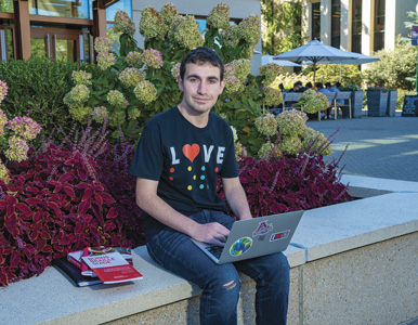 Harris Mowbray sits in front of the SIS buiilding with a laptop and wearing a shirt that reads "Love" in Braille