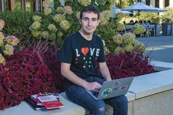 Harris Mowbray sits in front of the SIS buiilding with a laptop and wearing a shirt that reads "Love" in Braille