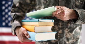 An American soldier holding a stack of schoolbooks, standing in front of an American flag