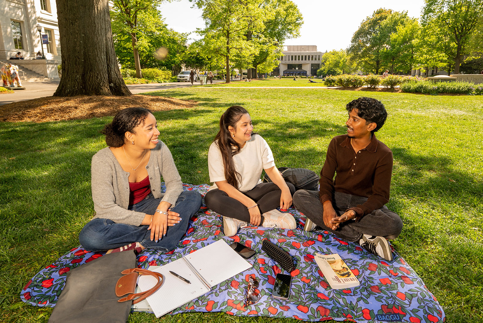 Three students sitting on a blanket talking to each other on the quad.