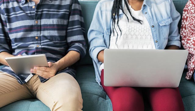 two students sit on a couch with a laptop and ipad in their hands