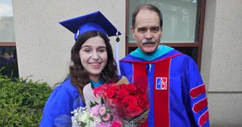 Tom and Ava outside SIS. Ava is dressed in graduation regalia and holding flowers. Tom is also in academic robes.