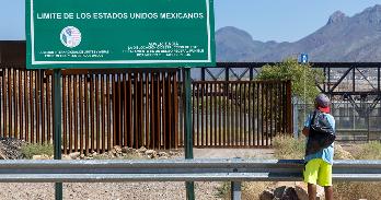 Migrant walks on road in front of sign for US border in Spanish