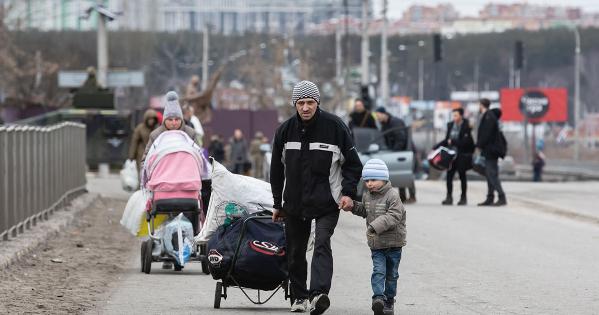 A man pulls luggage and holds a small boy's hand as they walk toward the camera. In the background, other refugees do similarly.