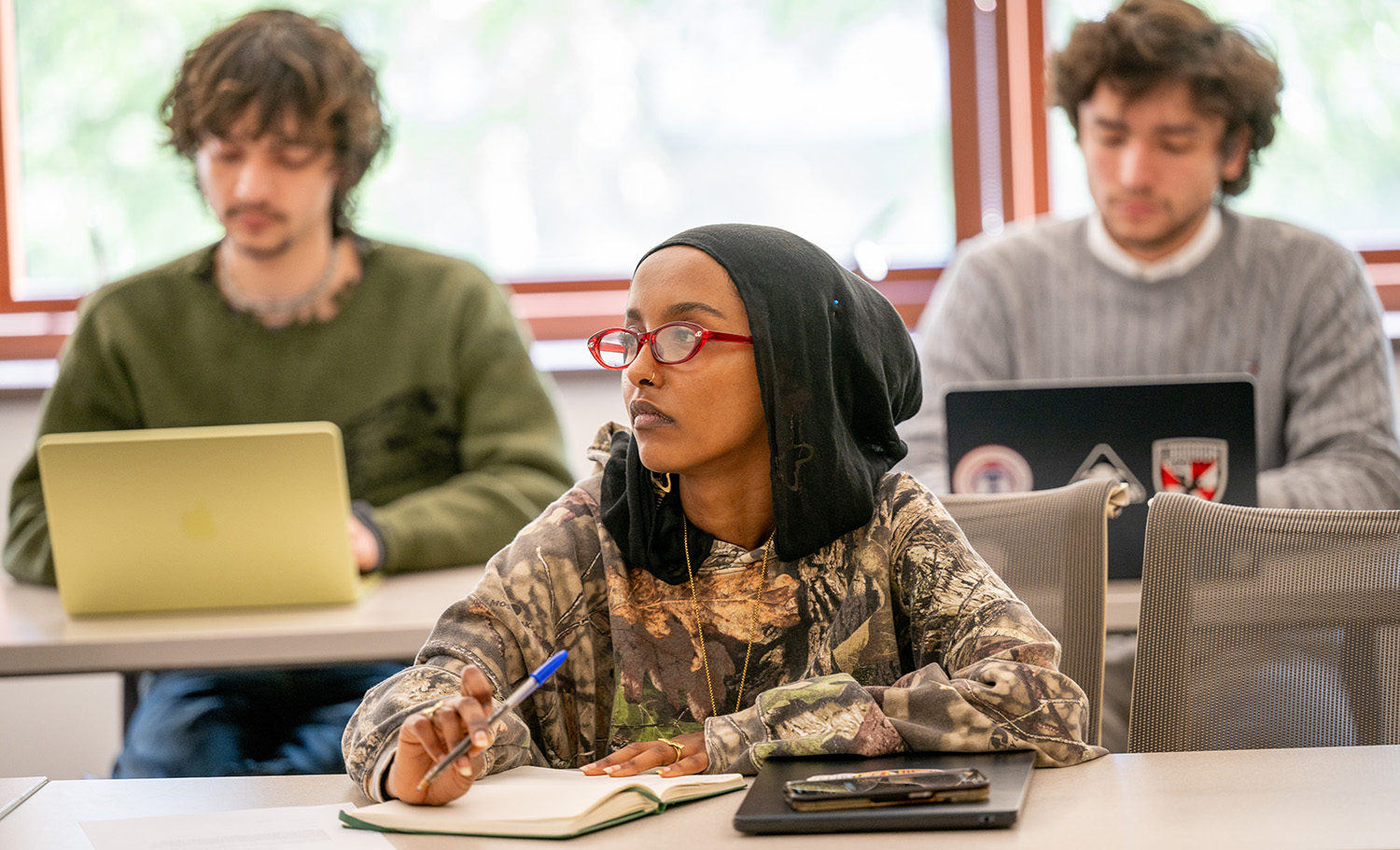 A young woman in glasses listens and takes notes during a class.