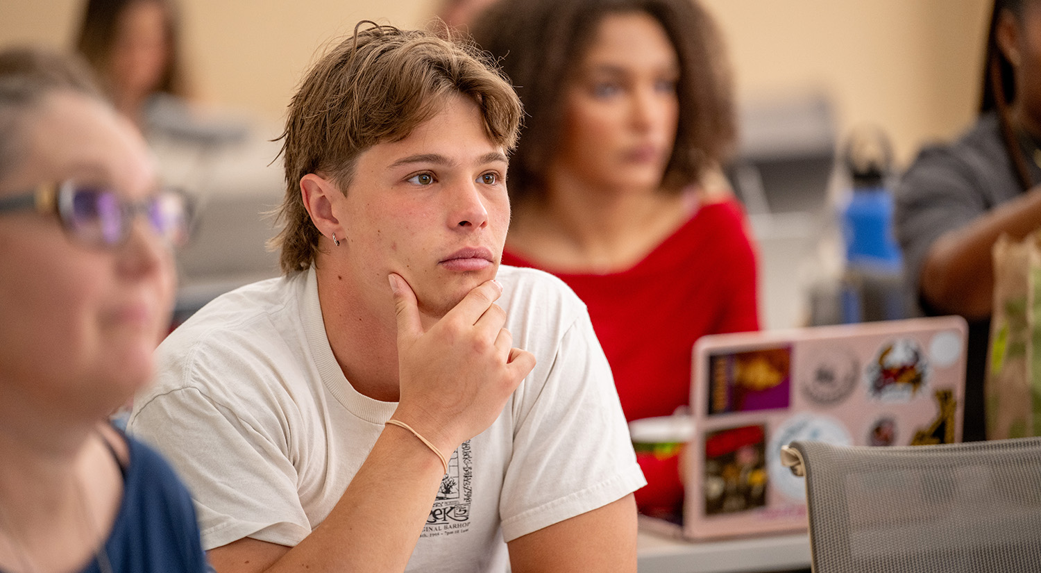 A student rests his chin on his hand at a classroom table as he listens intently.
