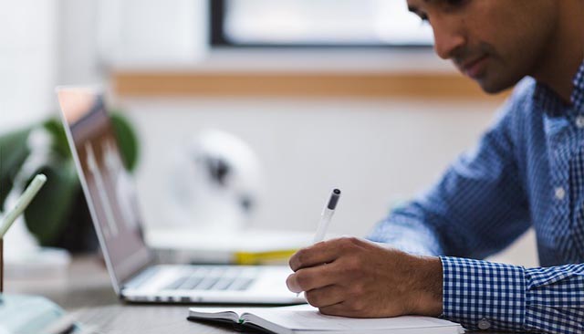 man sits at a laptop writing in a notepad