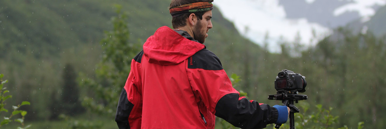 A man holds a camera on a steadycam with mountain scenery in the background.