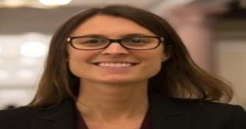 White woman with dark hair wearing a black shirt and square glasses stands before a tan, blurry background