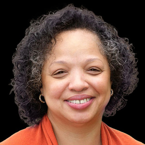 Vernice Miller-Travis, a middle age, woman of color wearing small hoop earrings and an orange sweater stands before a black background
