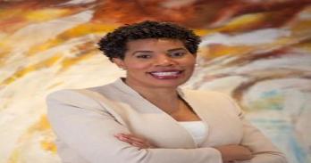 Margot Brown, a middle age, woman of color with short hair wearing a tan suit stands before a colorful background