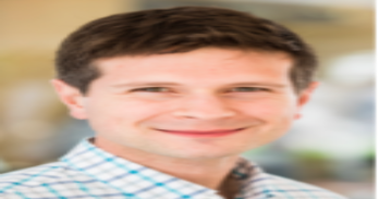 Joe Bonfiglio, a white man wearing a checked, collared shirt stands before blurry, tan background