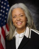 Robin Morris Collin, a middle age, woman of color wearing a collared shirt and sweater smiles at the camera while standing in front of a grey background with the American flag