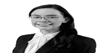 Daniela Stevens, a woman with long, dark hair and wearing glasses and a suit with a white blouse, stands before a white background.