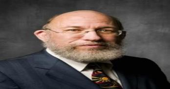 Scott Sklar, an older white man with no hair and a beard and wearing glasses and a suit and ties stands before a dark grey, blurred background