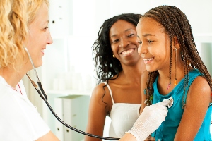 Doctor holds stethoscope to child's chest with woman behind child.