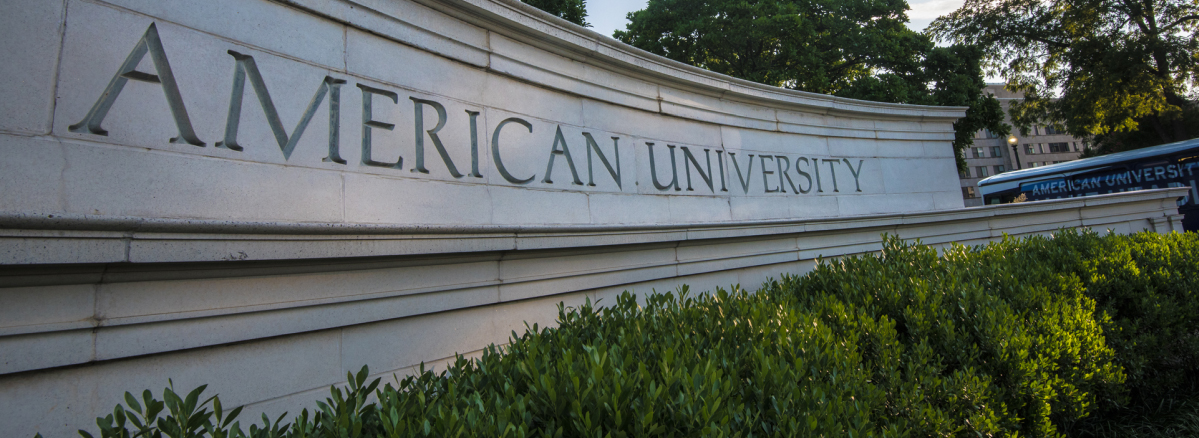 A stone sign reads American University. There are several green bushes underneath.
