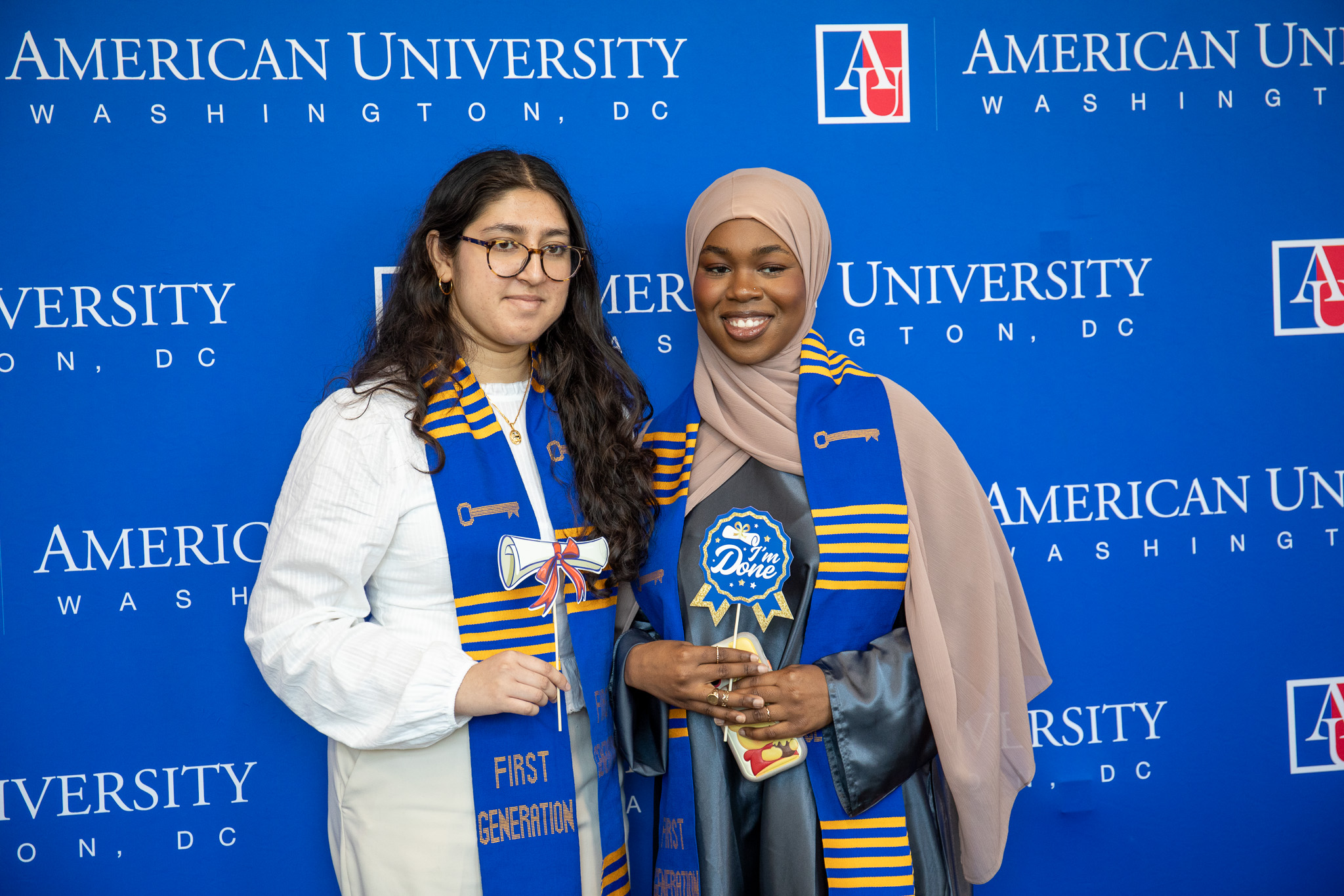 Two students wearing first generation stoles stand in front of an AU step and repeat and smile.