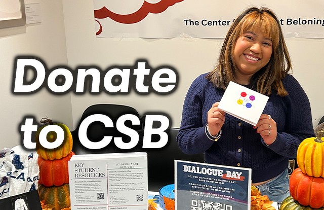 A student stands behind a table in the Center for Student Belonging, holding up a notecard. Text to the right of the student reads: Donate to CSB.