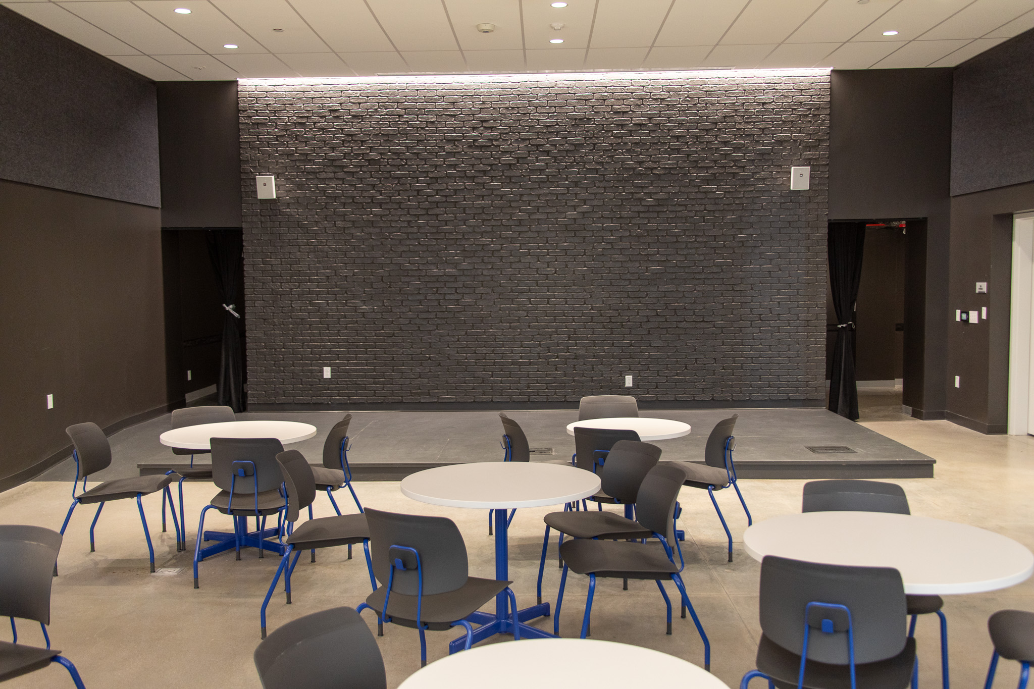 A blackbox-style theater and stage is at the back of a room. The foreground of the room is occupied by several tables with chairs tucked in.