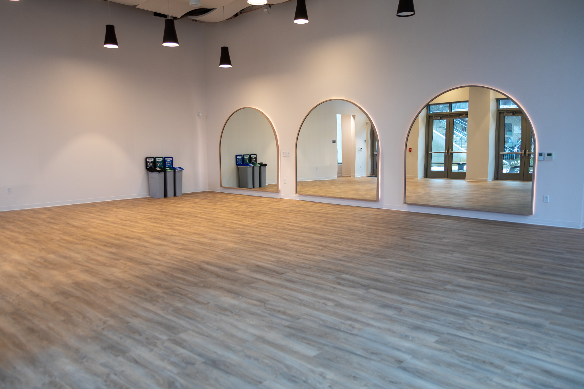 A bright empty room with wooden floors.  It resembles an empty yoga or dance studio, with three large mirrors on the wall.