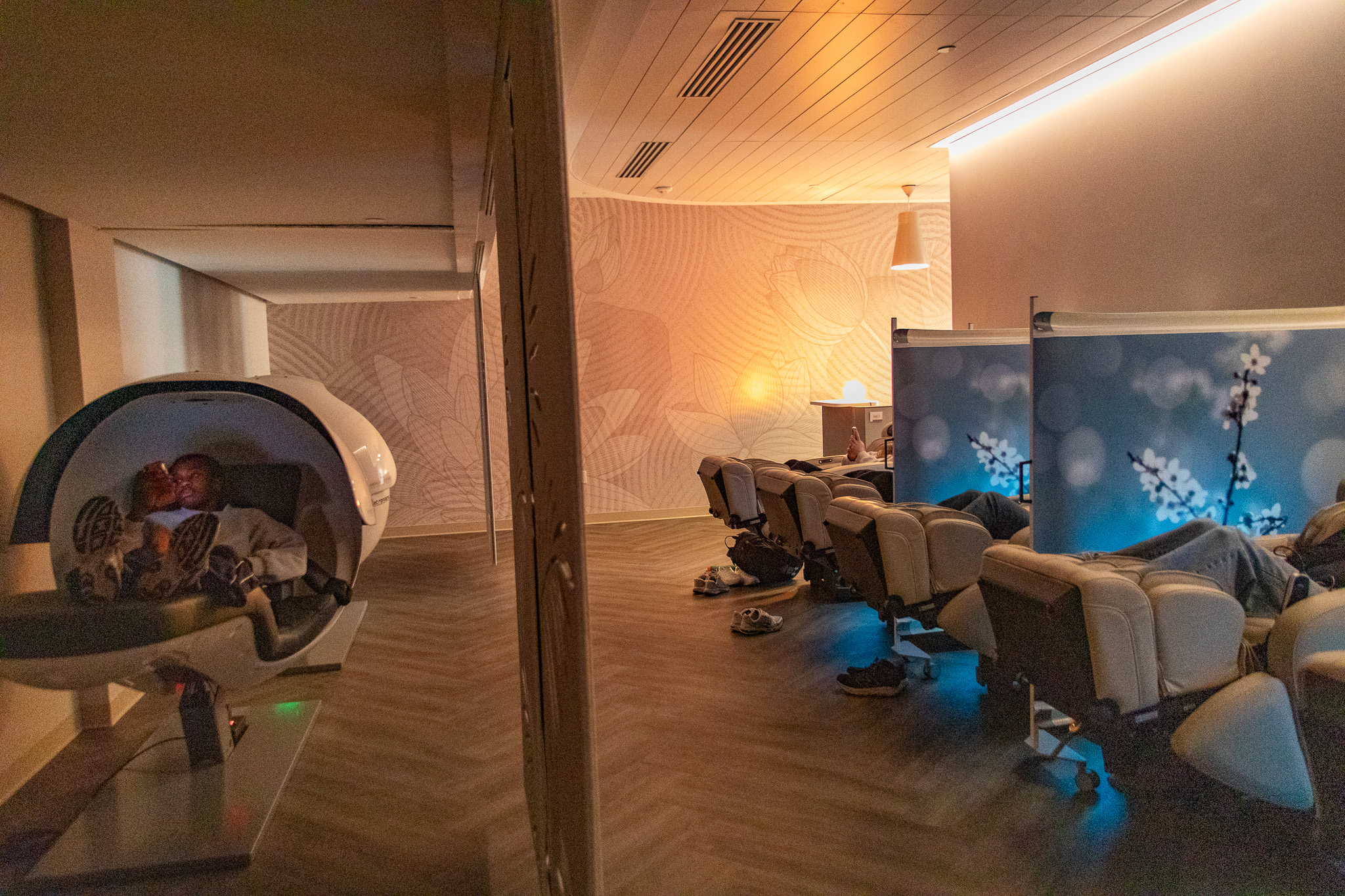 A dimly lit room with a student sitting in a sleeping pod to the right. On the left, students sitting in massage chairs are obscured by screen dividers.