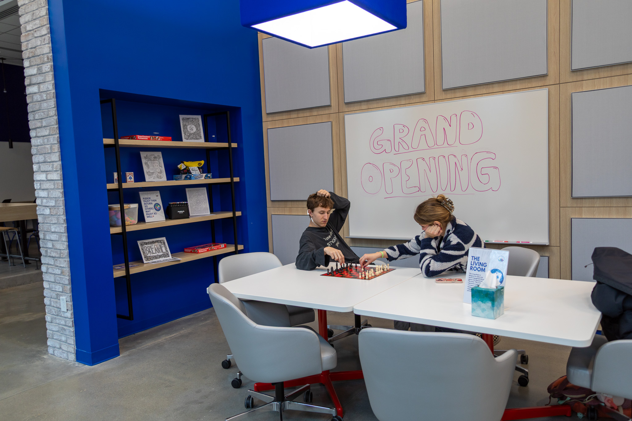 Two students sit at a table playing chess. A whiteboard behind them reads "Grand Opening." Nearby, a bookshelf built into the wall has several board games, coloring sheets, and other activity bins.