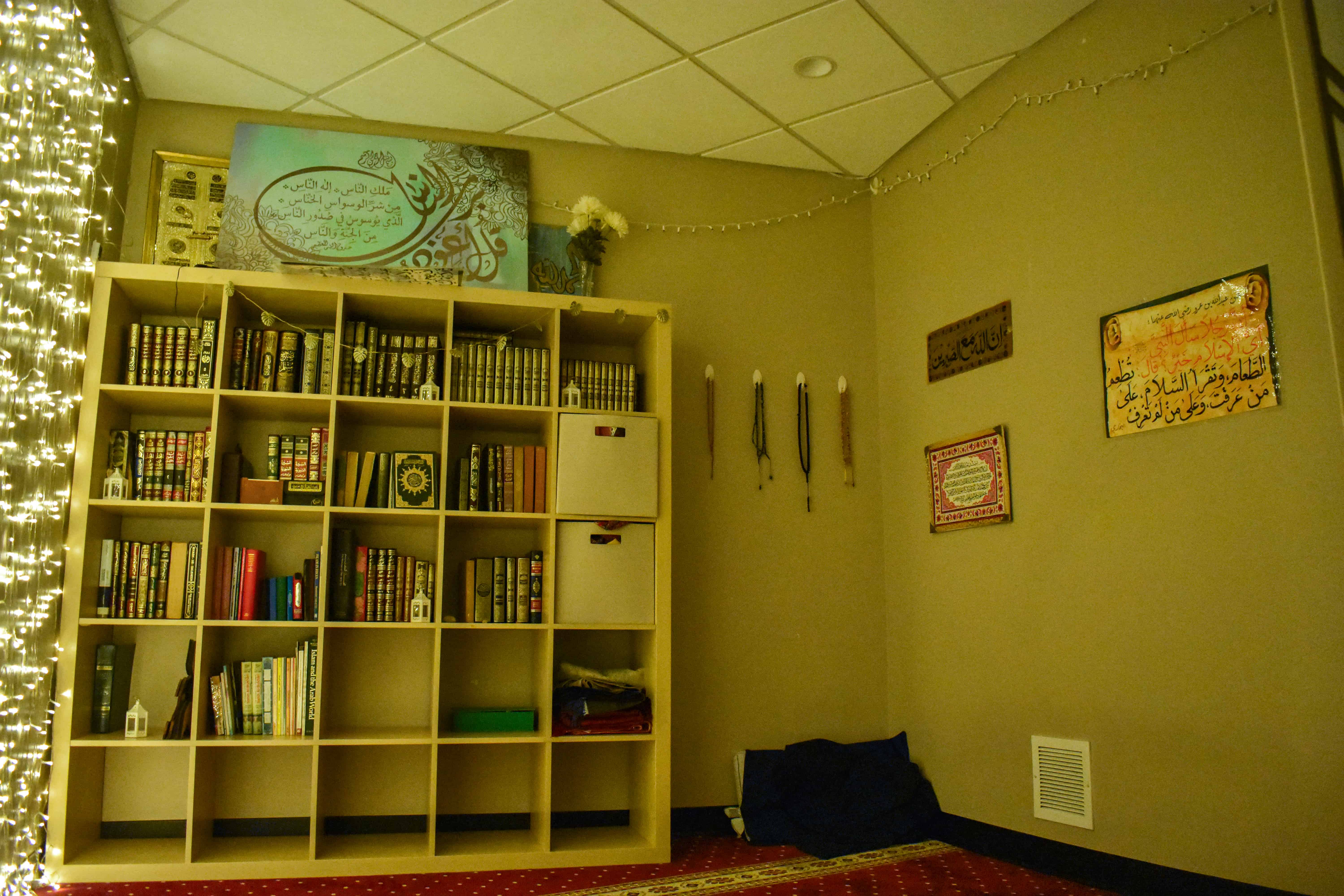 Muslim Prayer Room in Kay, showing a large bookshelf with lights besides it, as well as several wall hangings written in classical Arabic.