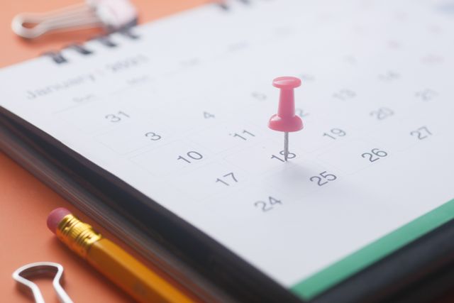 A close-up of a calendar page with a pink push pin, pencil, and binder clip on an orange background