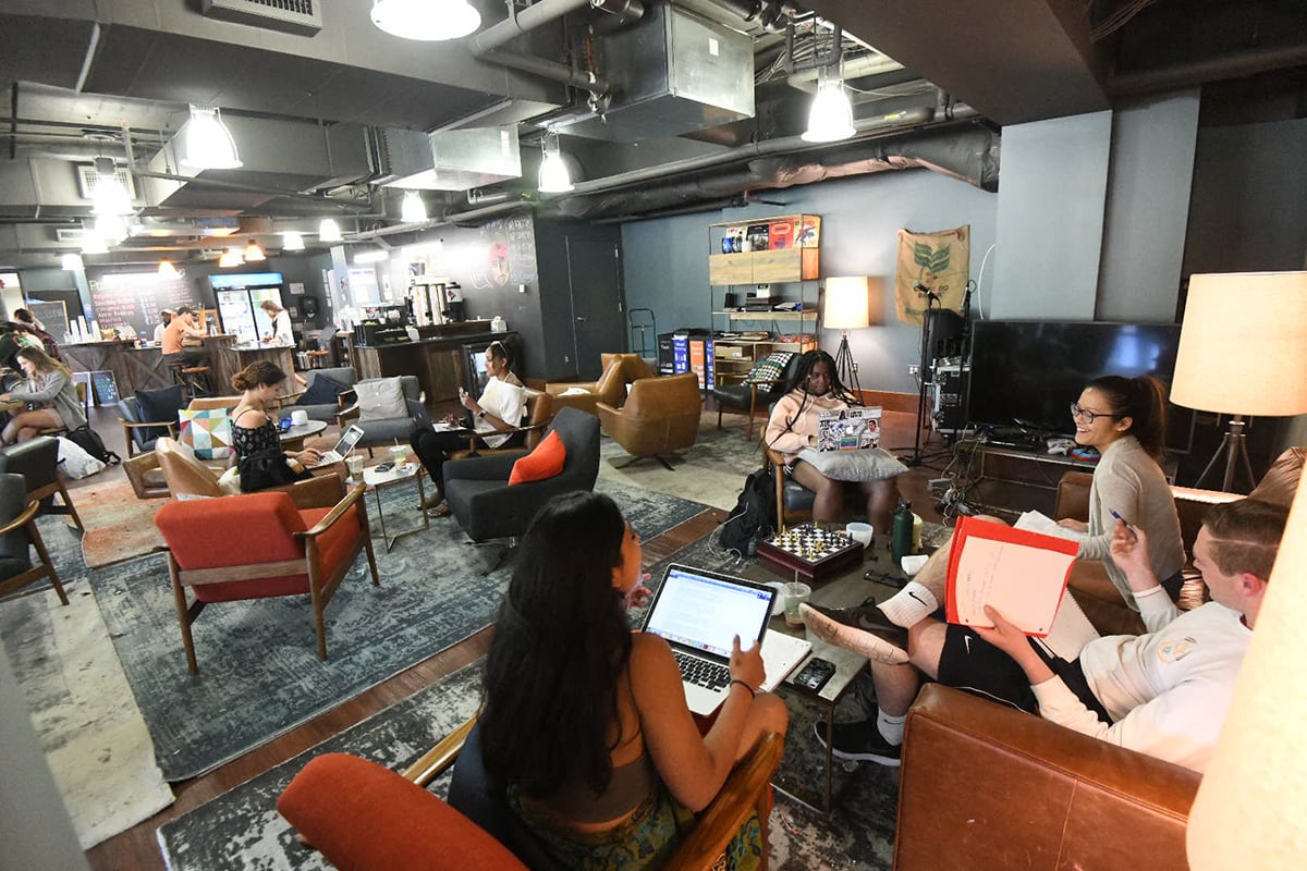 Students sit in the Bridge Cafe, working on their laptops in several orange chairs.