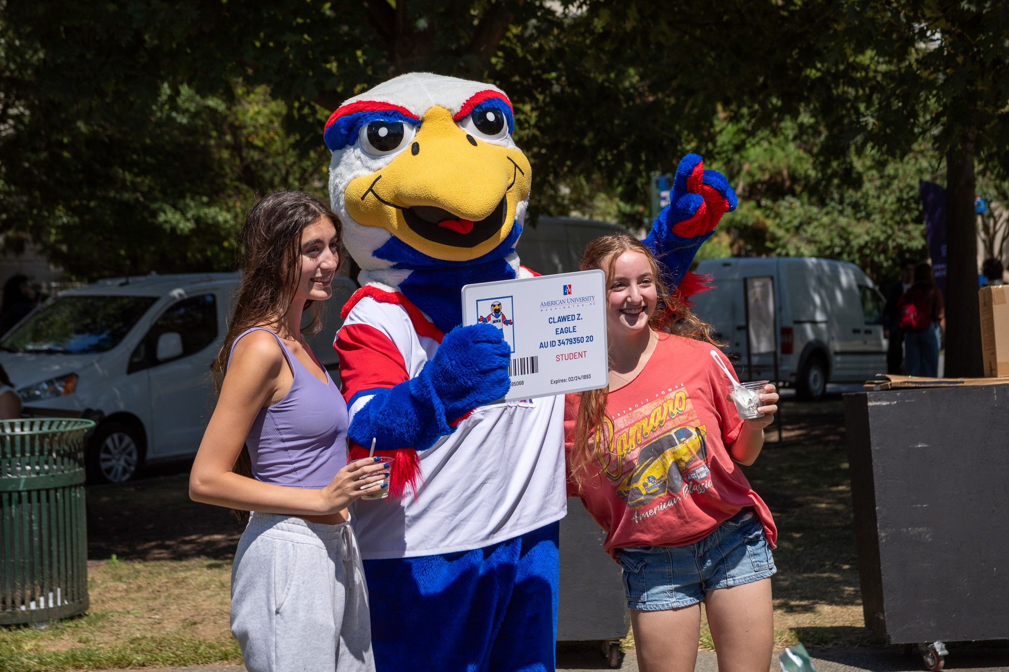 Two students pose with Clawed on the Quad, AU's mascot. Clawed holds a sign that looks like a student ID, while the two students hold cups with ice cream.