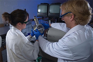 Two people in safety goggles and lab coats adjust a machine