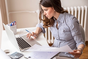 A young woman sits at a work desk in front of a laptop, taking notes