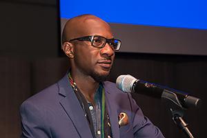 Man with glasses wearing with a blue sport jacket, in front of a microphone.