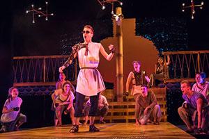 An actor dressed as an ancient greek peasant sings with nautical props. A group of similarly dressed actors kneel around him.