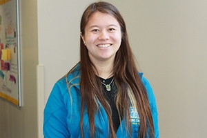 Kirsten Jay in a blue hoodie. Long brown hair and smiling in a hallway in MGC.